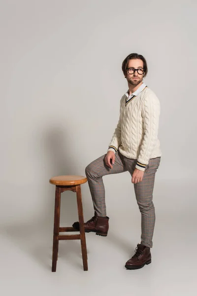 Модний чоловік в светрі і штанях дивиться далеко від високого, дерев'яний табурет на сірому — стокове фото