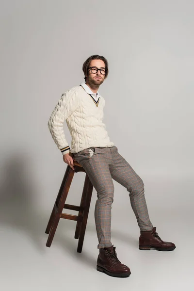 Young man in white sweater and pants sitting on high wooden stool on grey background - foto de stock