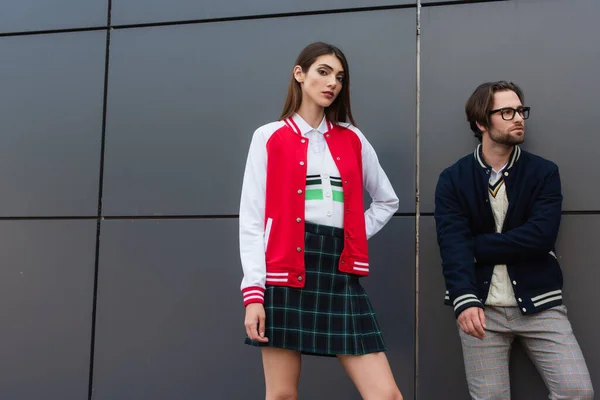 Stylish woman with hand on hip looking at camera near stylish man and grey wall - foto de stock