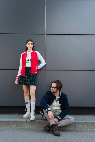 Young stylish woman looking at camera near trendy man sitting on border outdoors — Fotografia de Stock