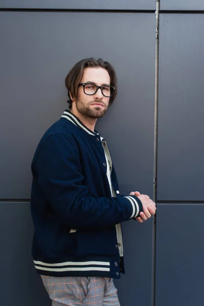 Trendiger Mann mit Brille und schicker Jacke blickt nahe grauer Wand in die Kamera — Stockfoto