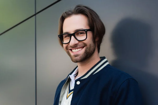Щасливий молодий чоловік в стильних окулярах стоїть біля сірої стіни, посміхаючись на камеру — стокове фото