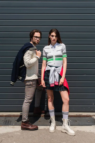 Молода пара в модному одязі дивиться на камеру біля сірої стіни — стокове фото