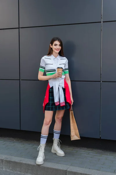 Trendy woman with coffee to go and shopping bags smiling near grey wall — Foto stock