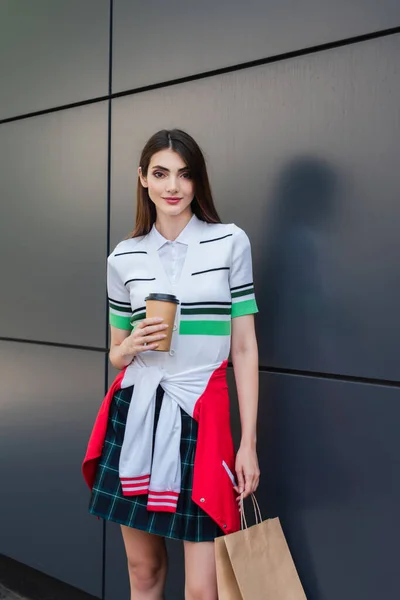 Positive woman in stylish clothes standing near grey wall with coffee to go and shopping bag — Photo de stock