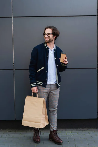 Cheerful and stylish man standing by grey wall with paper cup and shopping bags — Foto stock