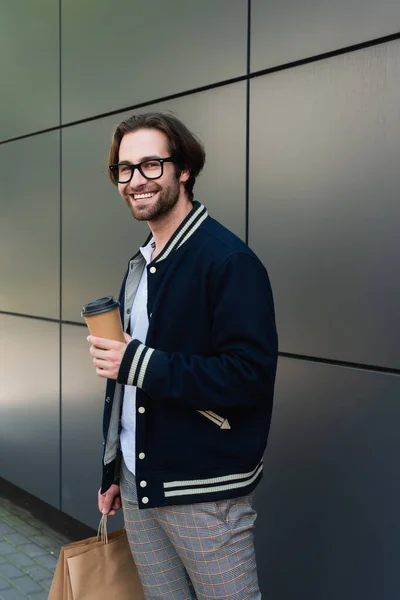 Joyful man in trendy jacket and eyeglasses holding takeaway drink near grey wall — Foto stock