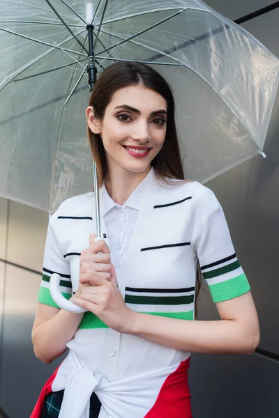 Joyful woman in striped polo t-shirt smiling at camera under transparent umbrella — Photo de stock