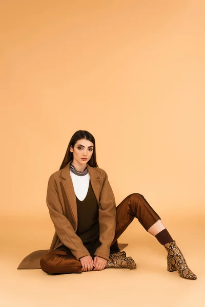 Pretty woman in trendy coat, satin pants and leather boots sitting on beige background — Stock Photo