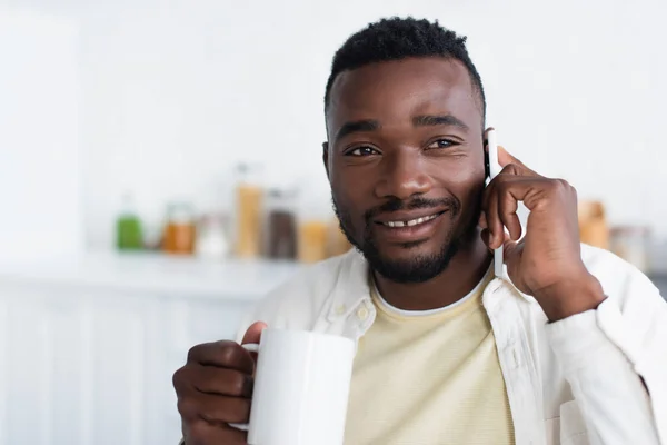 Feliz afro-americano homem segurando xícara de café e falando no celular — Fotografia de Stock