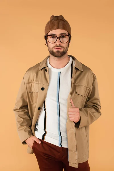 Young man in stylish eyeglasses, jacket and beanie posing with hand in pocket isolated on beige — Photo de stock