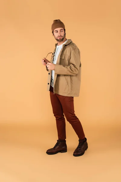 Full length view of trendy man looking back while holding eyeglasses on beige — Foto stock