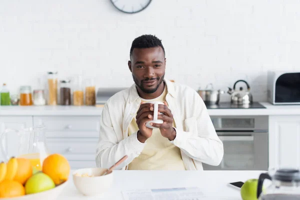 Hombre afroamericano feliz sosteniendo taza de café cerca del desayuno en la mesa - foto de stock
