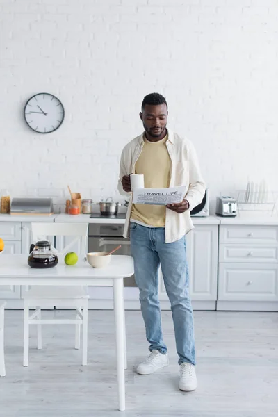 Joven afroamericano hombre sosteniendo taza de café y leyendo viaje vida periódico en cocina - foto de stock