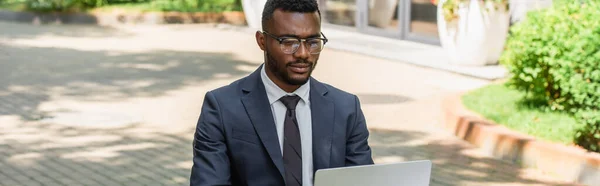 Joven hombre de negocios afroamericano en gafas usando el ordenador portátil fuera, pancarta - foto de stock
