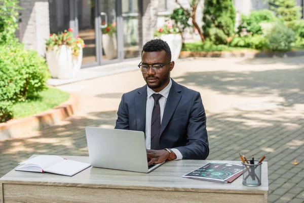 Joven hombre de negocios afroamericano en gafas usando el ordenador portátil fuera - foto de stock