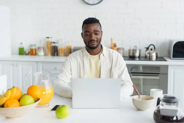 Felice giovane uomo africano americano utilizzando il computer portatile in cucina — Foto stock