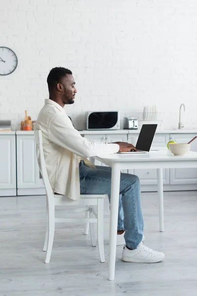 Side view of young african american man using laptop with blank screen in kitchen — Stock Photo