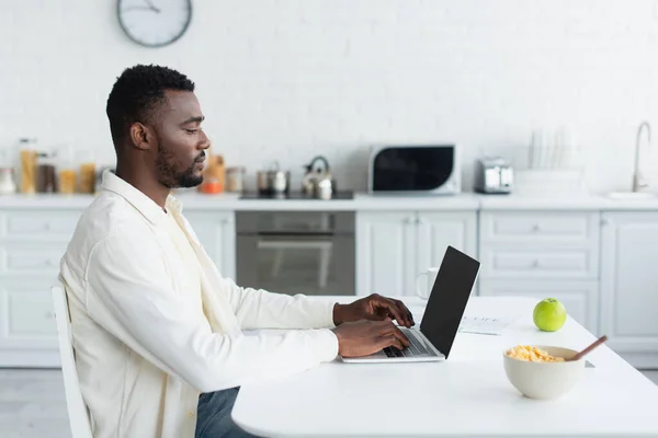 Side view of young african american freelancer using laptop with blank screen in kitchen — Stock Photo