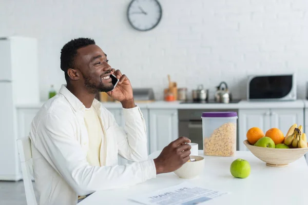 Hombre afroamericano feliz hablando en el teléfono inteligente mientras desayuna - foto de stock