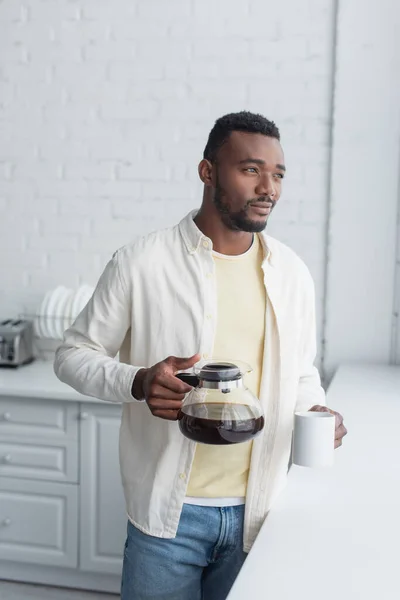 Молодой африканский американец держит кофейник и кружку на кухне — стоковое фото