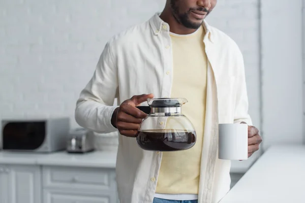 Vista cortada do jovem afro-americano segurando panela de café e caneca na cozinha — Fotografia de Stock