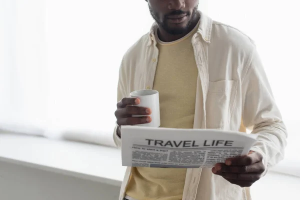 Vista recortada del hombre afroamericano sosteniendo una taza de café y leyendo el periódico de la vida de viaje - foto de stock