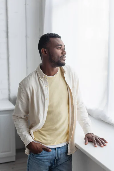 Nachdenklicher afrikanisch-amerikanischer Mann im Hemd, der mit der Hand in der Tasche am Fenstersims steht — Stockfoto