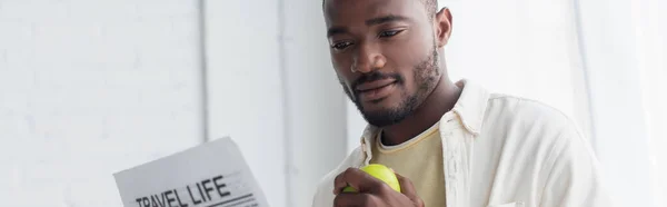 Hombre afroamericano sosteniendo manzana y leyendo periódico de la vida de viaje, pancarta - foto de stock