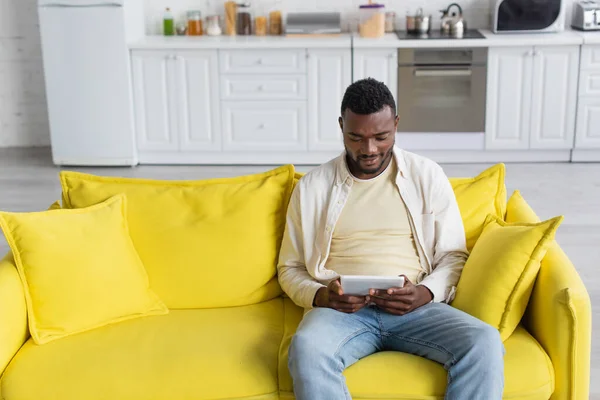 Hombre afroamericano sonriente usando tableta digital mientras está sentado en el sofá - foto de stock