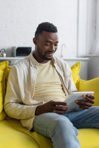 Fröhlicher afrikanisch-amerikanischer Mann nutzt digitales Tablet während er auf der Couch sitzt — Stockfoto