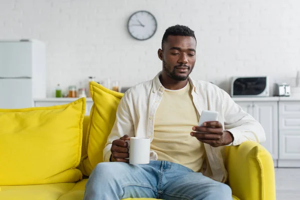 Молодой африканский американец с помощью смартфона, сидя на диване с чашкой — стоковое фото