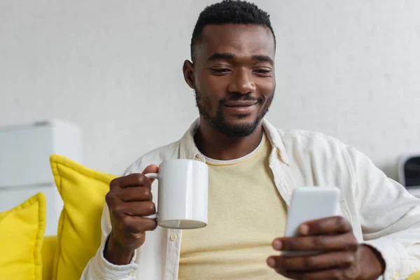 Sorridente giovane uomo africano americano utilizzando il cellulare e tenendo in mano la tazza — Foto stock