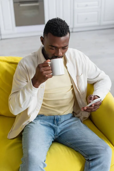 Vista de ángulo alto de hombre afroamericano joven usando el teléfono celular y beber café mientras está sentado en el sofá - foto de stock