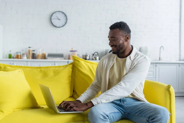 Alegre afroamericano freelancer escribiendo en el ordenador portátil mientras está sentado en el sofá amarillo - foto de stock