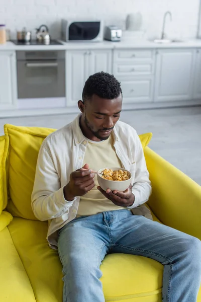 Joven afroamericano hombre mirando tazón con hojuelas de maíz y sentado en sofá - foto de stock