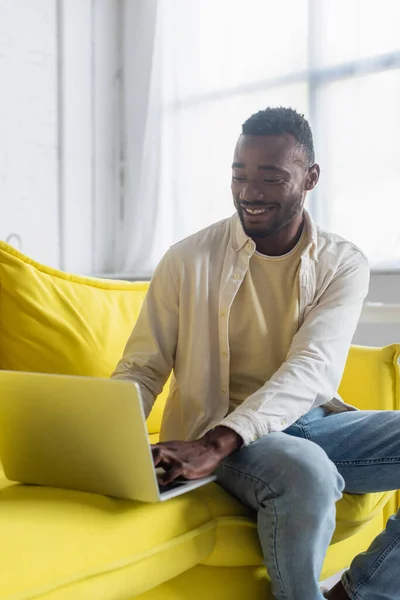 Glückliche junge afrikanisch-amerikanische Freiberuflerin tippt auf dem Laptop, während sie auf der gelben Couch sitzt — Stockfoto