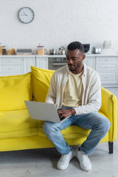 Joven afroamericano freelancer escribiendo en el ordenador portátil mientras está sentado en el sofá amarillo - foto de stock