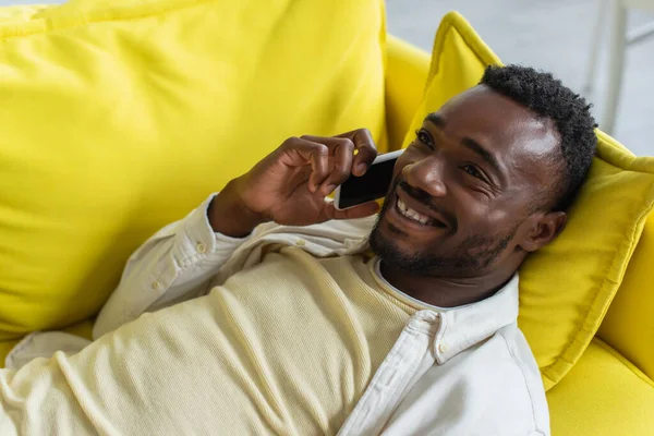 Glücklicher afrikanisch-amerikanischer Mann im Smartphone-Gespräch auf gelbem Sofa liegend — Stockfoto