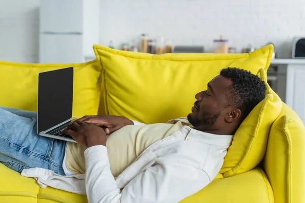 Afrikanisch-amerikanischer Freiberufler tippt auf Laptop, während er auf gelbem Sofa liegt — Stockfoto