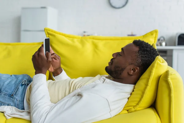 Afrikanisch-amerikanischer Mann benutzt Smartphone während er auf gelbem Sofa liegt — Stockfoto