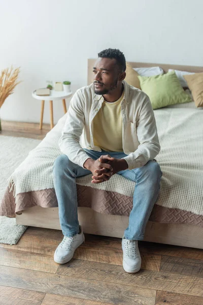 Задумчивый молодой африканский американец с сжатыми руками, сидящий на кровати — стоковое фото