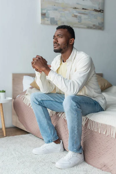 Вдумчивый молодой африканский американец с сжатыми руками, сидящий на кровати — стоковое фото