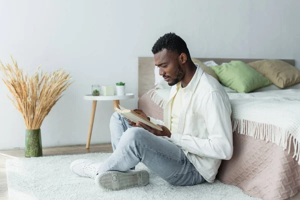 Junger afrikanisch-amerikanischer Mann liest Buch, während er mit überkreuzten Beinen in Bettnähe sitzt — Stockfoto