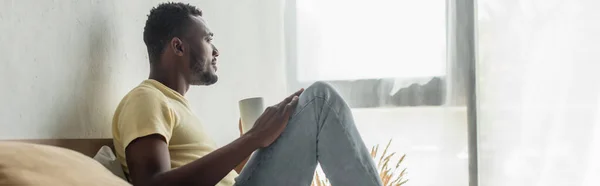 Vista lateral del hombre afroamericano sosteniendo taza de café en la cama, pancarta - foto de stock