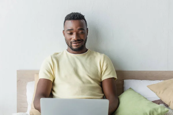 Alegre afroamericano freelancer utilizando ordenador portátil en el dormitorio - foto de stock