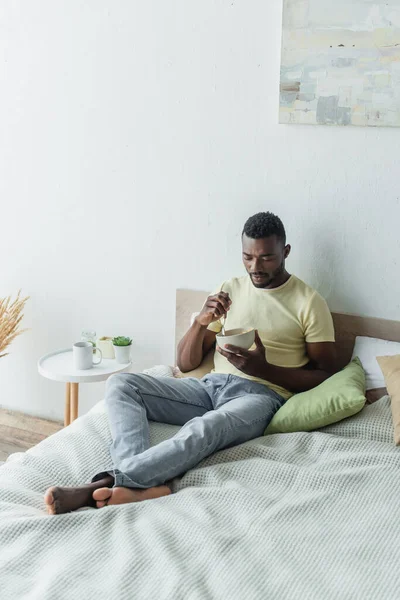 Barefoot african american man looking at corn flakes in bowl while chilling in bed — Stock Photo