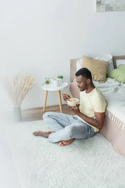Homem americano africano sentado com pernas cruzadas perto da cama enquanto toma café da manhã — Fotografia de Stock