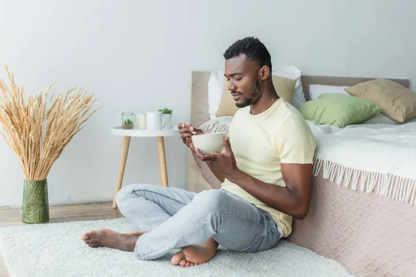 Homme afro-américain barbu assis avec les jambes croisées près du lit tout en prenant le petit déjeuner — Photo de stock