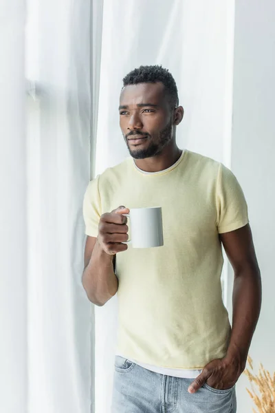 Barbudo afroamericano hombre sosteniendo taza con té y de pie con la mano en el bolsillo - foto de stock